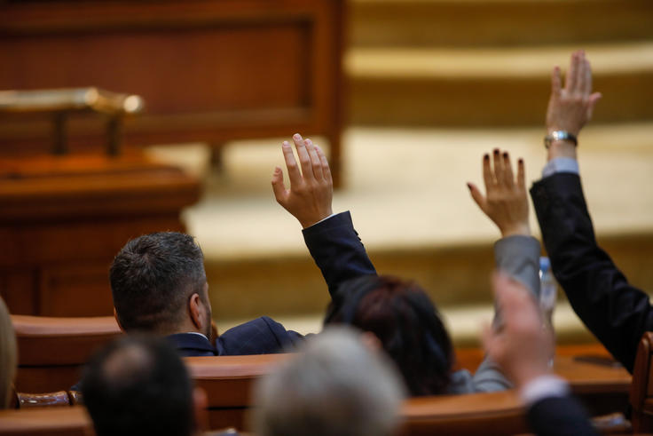 People in a meeting raising hands to vote, seated in a wooden chamber setting.