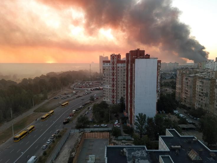 Smoke rises from buildings in a cityscape at sunset, with orange skies and a road lined with yellow buses.