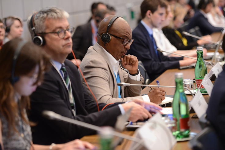 Conference attendees seated, wearing headsets, taking notes at a long table.