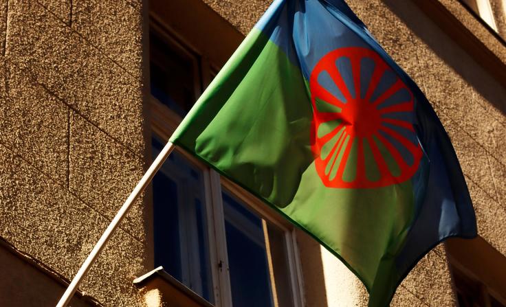 Romani flag with blue and green halves, red spoked wheel, waving on a building.
