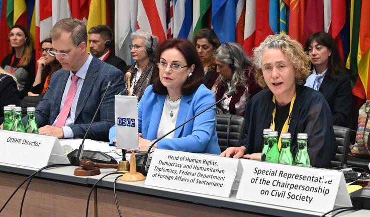 Three people sit at a conference table with microphones in front of national flags.