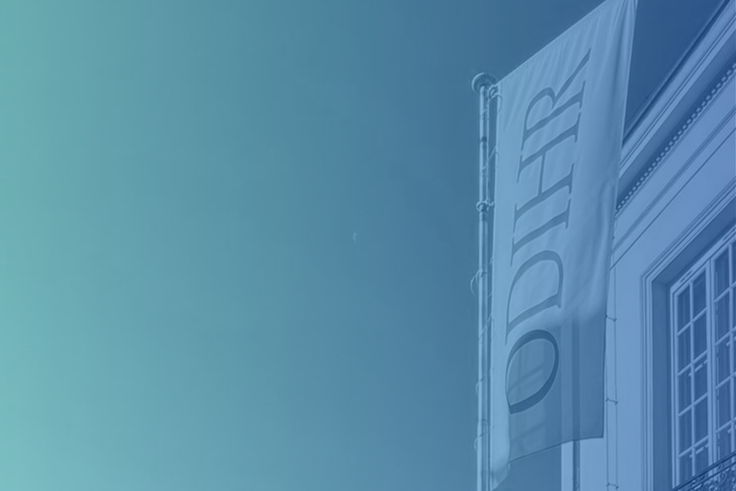 A flag with "ODIHR" on a building facade against a blue gradient sky.