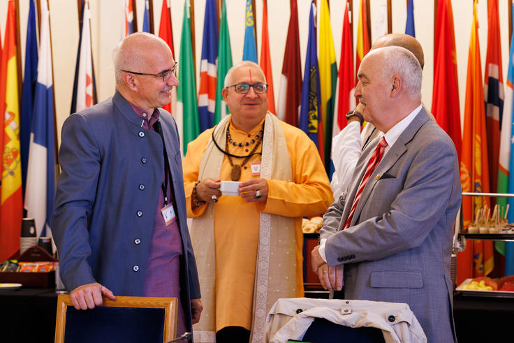 Three men in conversation at an event, with colorful flags in the background.