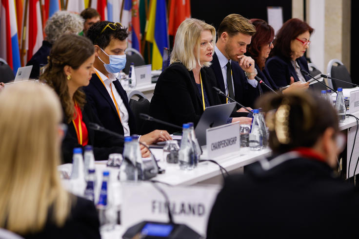 Conference attendees seated at a table with laptops, diverse flags in the background.