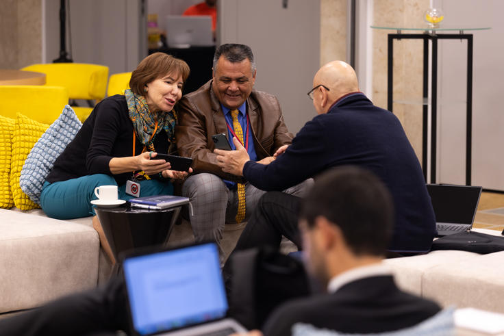 Three people engaged in a discussion on a sofa, smiling. A person in the foreground uses a laptop.