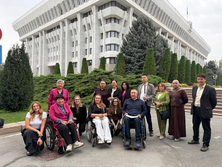 Diverse group of people, some in wheelchairs, smiling in front of a large government building with greenery.