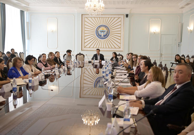 People sitting around a large conference table in a formal meeting room with chandeliers.