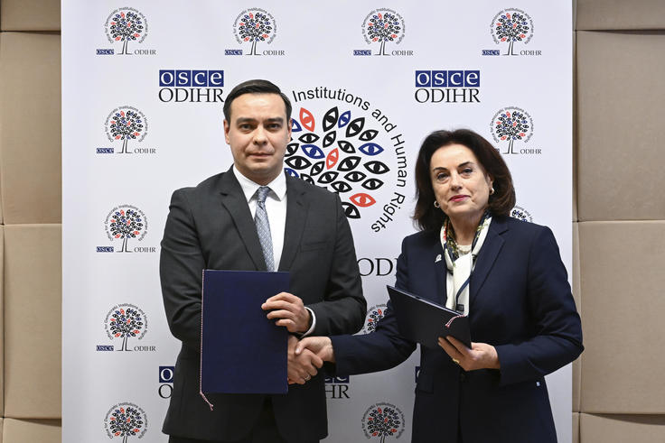 Two people in formal attire shake hands while holding folders, standing in front of a branded backdrop.