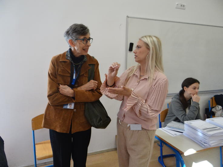 Two women in conversation in a classroom; one gesturing, others seated at desks with papers.