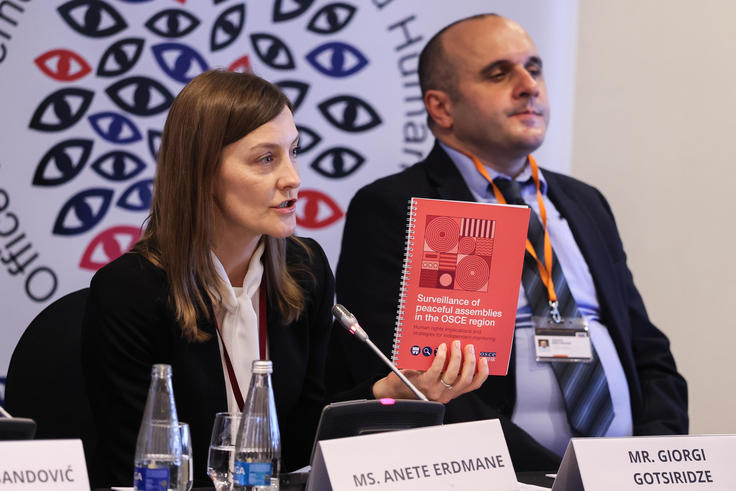 A woman speaks at a conference, holding a red book, seated next to a man. Logo with eyes in the background.
