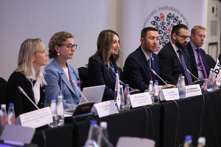 Panel of six people seated at a conference table with microphones and laptops.