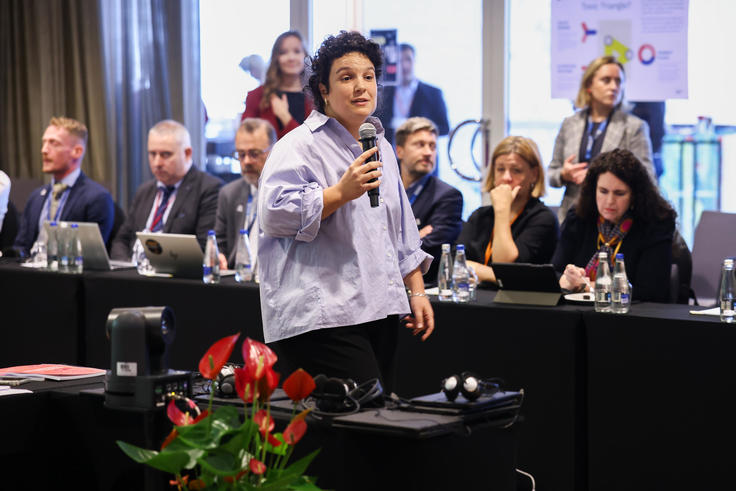 A person speaks into a microphone in a conference room with an attentive audience seated at tables.
