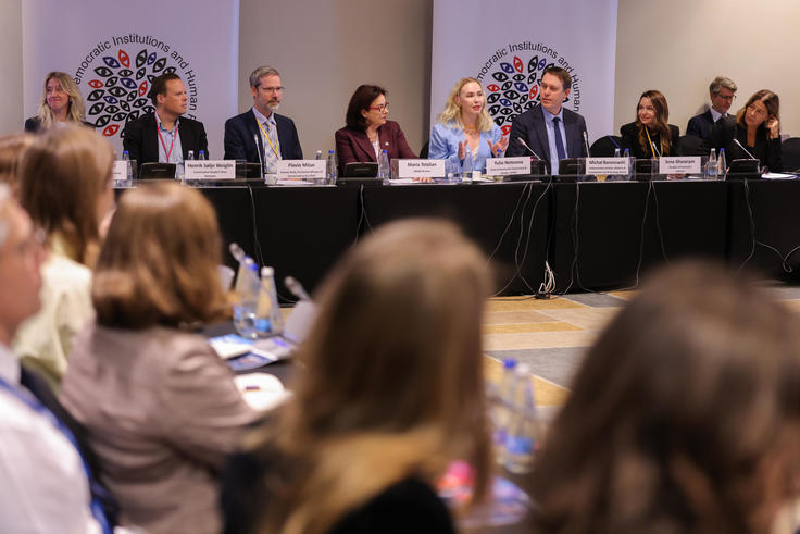 Panel discussion with speakers seated at a long table, audience in foreground, logos on banners behind.