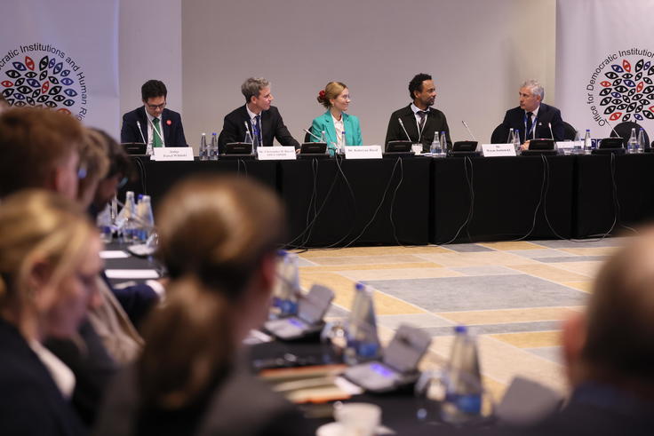 Conference panel discussion with six speakers seated at a table, blurred audience in the foreground.