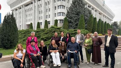 Diverse group of people, some in wheelchairs, smiling in front of a large government building with greenery.