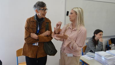 Two women in conversation in a classroom; one gesturing, others seated at desks with papers.