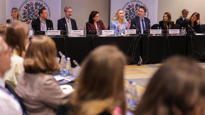 Panel discussion with speakers seated at a long table, audience in foreground, logos on banners behind.