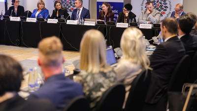 Conference panel discussion with attendees seated around a table, focusing on a row of speakers addressing the audience.