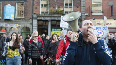 Protester speaking into a megaphone at a street demonstration, surrounded by a crowd with signs in an urban setting.