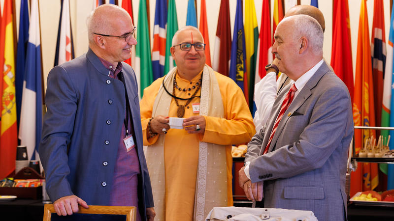 Three men in conversation at an event, with colorful flags in the background.
