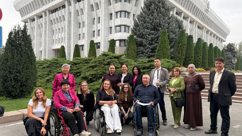 Diverse group of people, some in wheelchairs, smiling in front of a large government building with greenery.