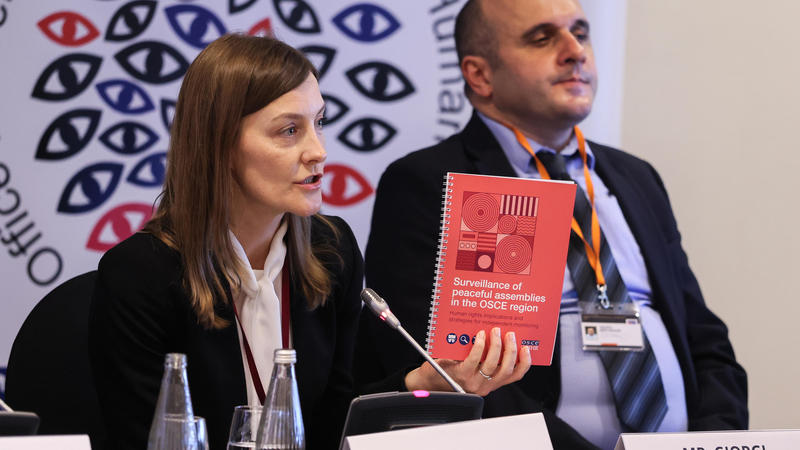 A woman speaks at a conference, holding a red book, seated next to a man. Logo with eyes in the background.
