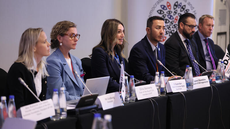 Panel of six people seated at a conference table with microphones and laptops.