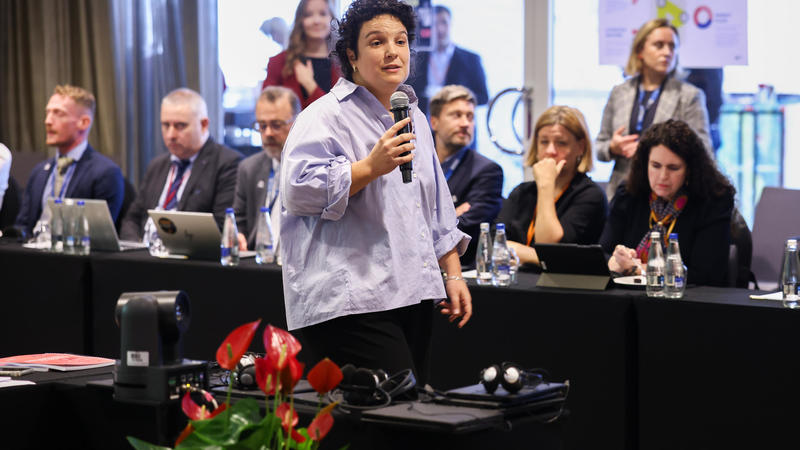 A person speaks into a microphone in a conference room with an attentive audience seated at tables.