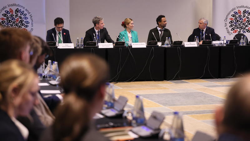 Conference panel discussion with six speakers seated at a table, blurred audience in the foreground.